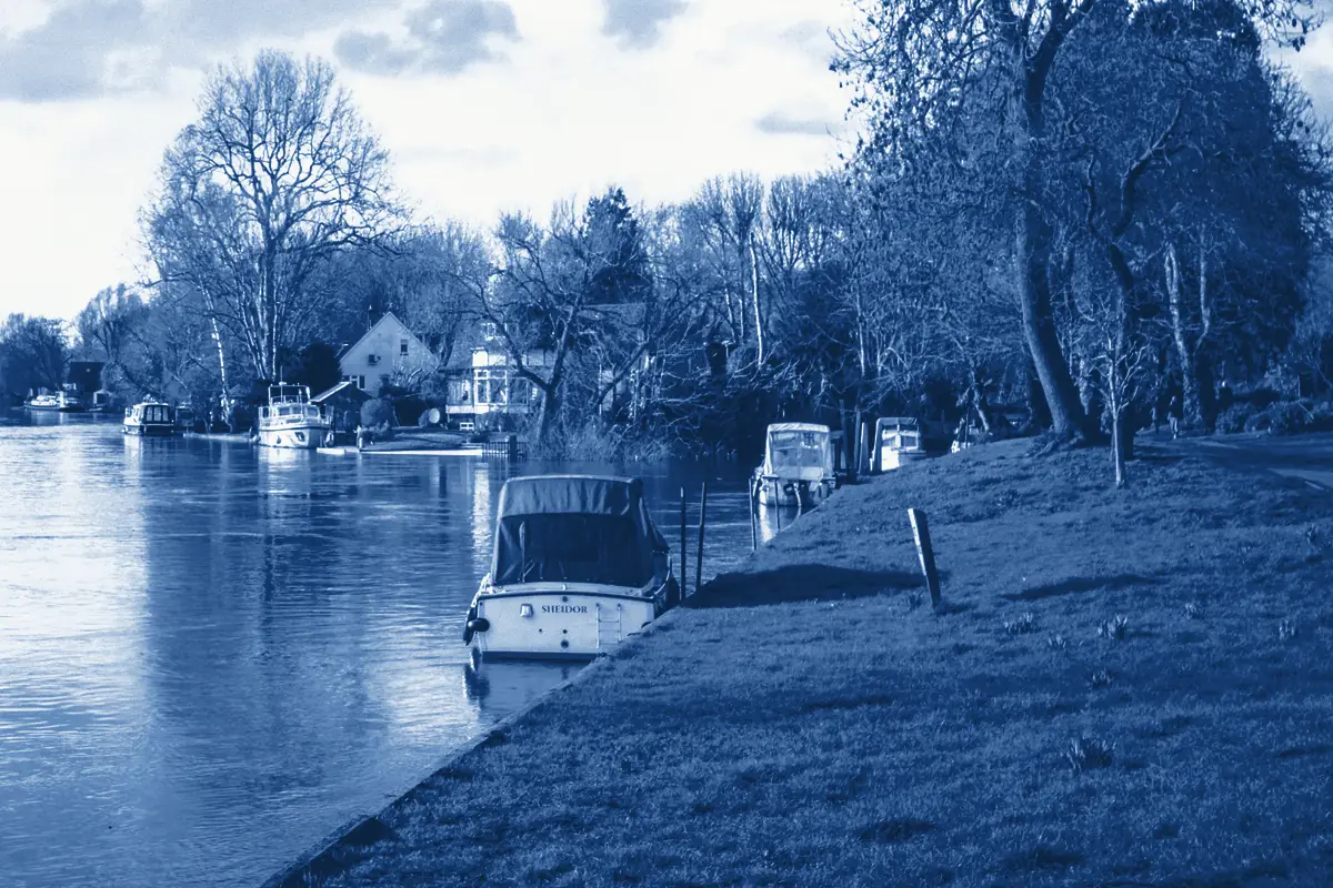 A covered boat moored by a grass bank on the Thames in winter, with houses and bare trees beyond, printed in Prussian blue.