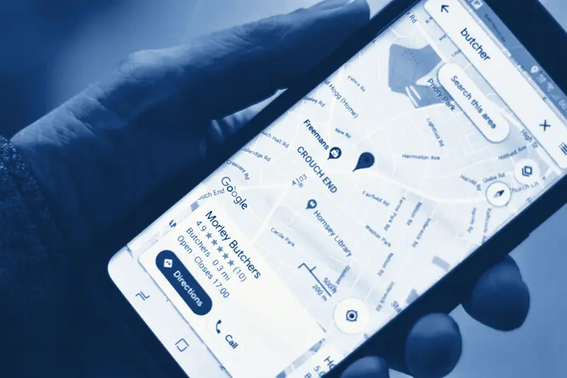 A hand holding a phone with Google Maps open on a search for a butcher, a shop’s listing card showing its stars, opening hours and buttons for directions and calling, printed in Prussian blue.