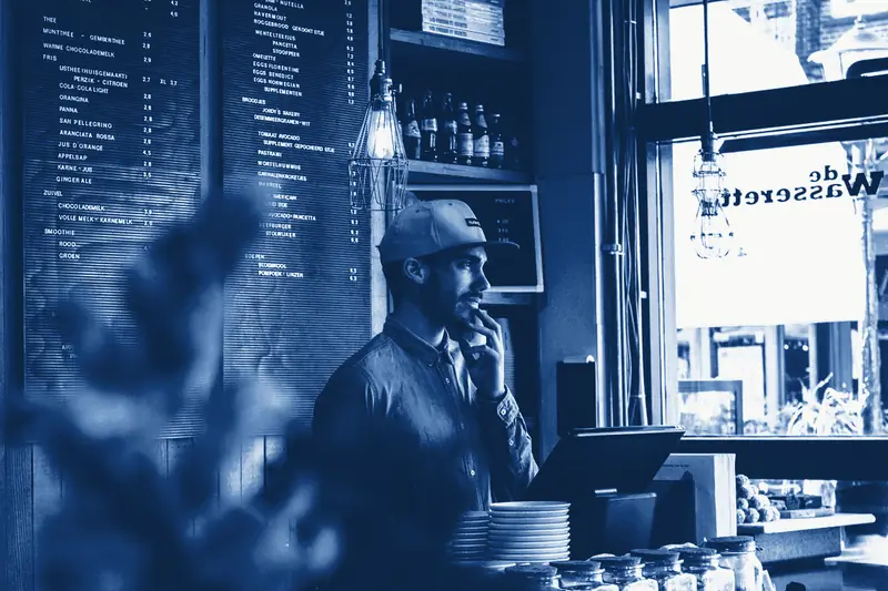 A caf&eacute; owner in a cap behind the counter, under the menu boards, printed in Prussian blue.