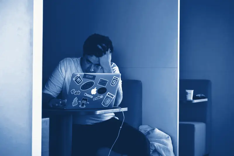 A man with his head in his hand, sitting at a laptop covered in stickers, printed in Prussian blue.