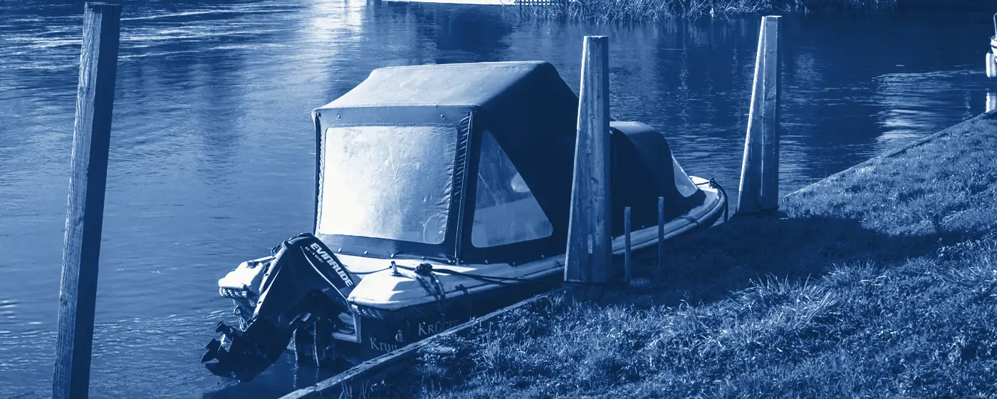 A covered motorboat tied to wooden mooring posts by a grass bank on the Thames, printed in Prussian blue.